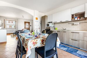 A modern kitchen with white cabinets, a microwave, and a sink. There is a dining area with a table set for four, chairs, and a blue rug. - GuestHost - Sweet Home in Portoscuso! (Portoscuso)