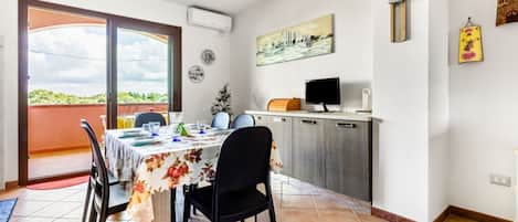 Uma sala de jantar com mesa posta para quatro pessoas, uma cozinha com geladeira e micro-ondas, e uma porta de vidro deslizante que dá para uma varanda.