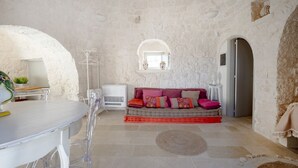 A room with a stone wall, a sofa with colorful cushions, a round rug, and a white table with chairs. - A charming trulli complex wit pool and 2000sqm of land planted with olive trees; it hosts 6 guests with 3 independent sleeping areas and 3 bathrooms. Located just 3 km from Alberobello and midway between the Adriatic and Ionian coasts, in the countryside (Martina Franca)