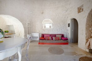 Ein Zimmer mit Steinwand, einer Couch mit bunten Kissen, einem runden Teppich und einem weißen Tisch mit Stühlen.