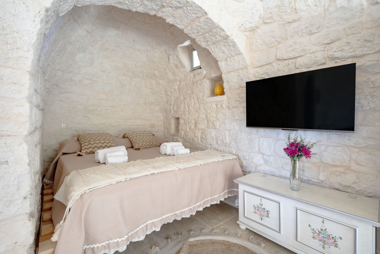 Ein Schlafzimmer mit Steinwand, einem Bett mit beigefarbenem Bettbezug, einem Flachbildfernseher auf einer weißen Kommode und einer Vase mit Blumen.