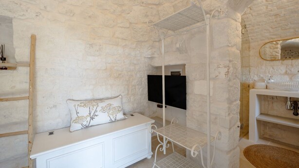 A room with a stone wall, a white bench with a decorative pillow, a white metal bed frame, a mounted TV, and a white sink with a mirror. - A charming trulli complex wit pool and 2000sqm of land planted with olive trees; it hosts 6 guests with 3 independent sleeping areas and 3 bathrooms. Located just 3 km from Alberobello and midway between the Adriatic and Ionian coasts, in the countryside (Martina Franca)