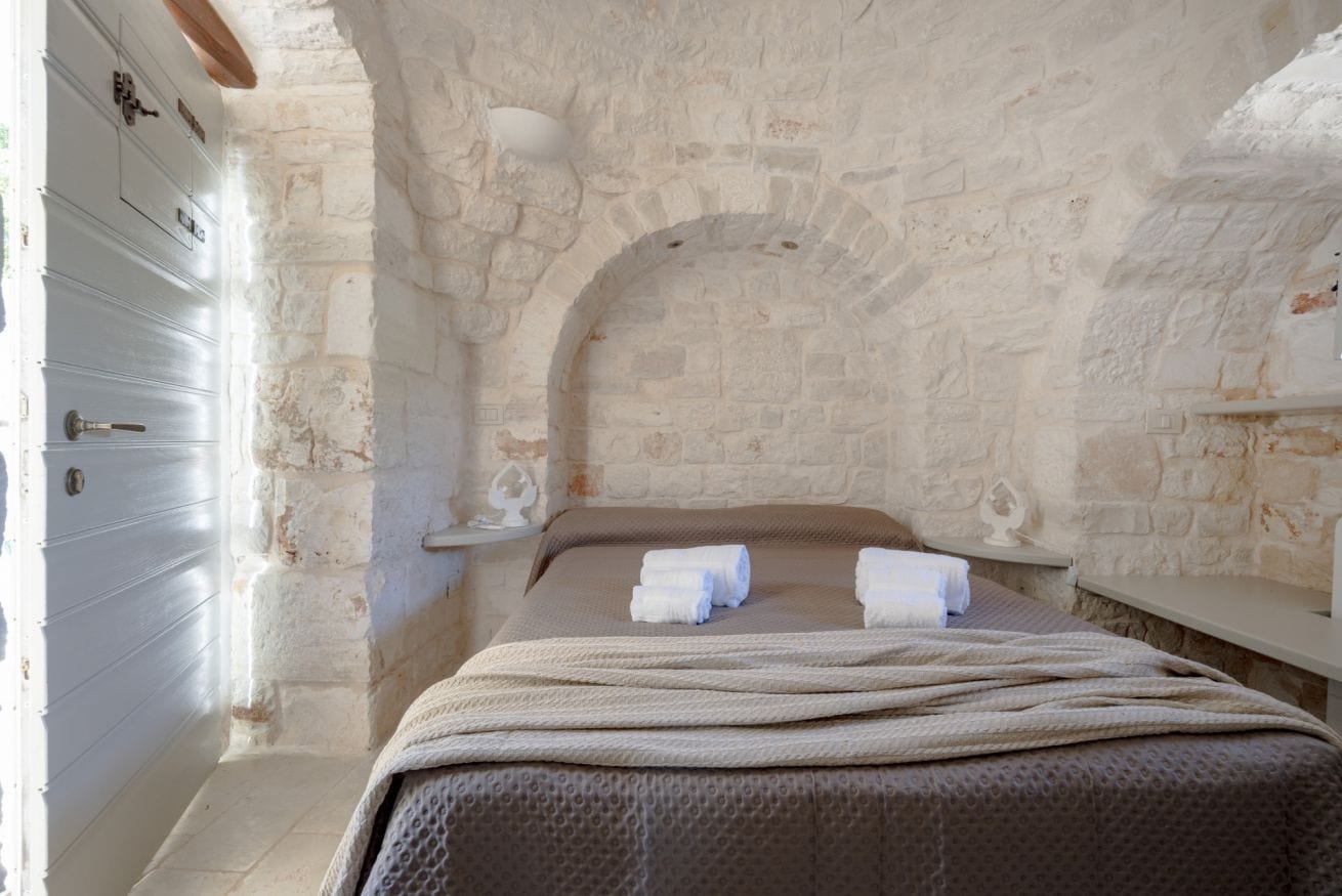 Une pièce avec un mur en pierre, un lit recouvert d’une couverture marron et une commode blanche à tiroirs.