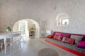 Ein Zimmer mit Steinsturz, einem weißen Sofa mit bunten Kissen, einem Esstisch mit Stühlen und einem kleinen Fenster.