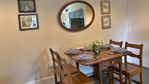 A dining area with a wooden table set for four, surrounded by chairs. There are framed pictures on the wall, a round mirror above the table, and a light fixture above. - Charming Snowdonia Cottage for 4 (Betws-Y-Coed)