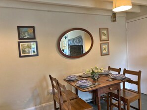 A dining area with a wooden table set for four, surrounded by chairs. There are framed pictures on the wall, a round mirror above the table, and a light fixture above. - Charming Snowdonia Cottage for 4 (Betws-Y-Coed)