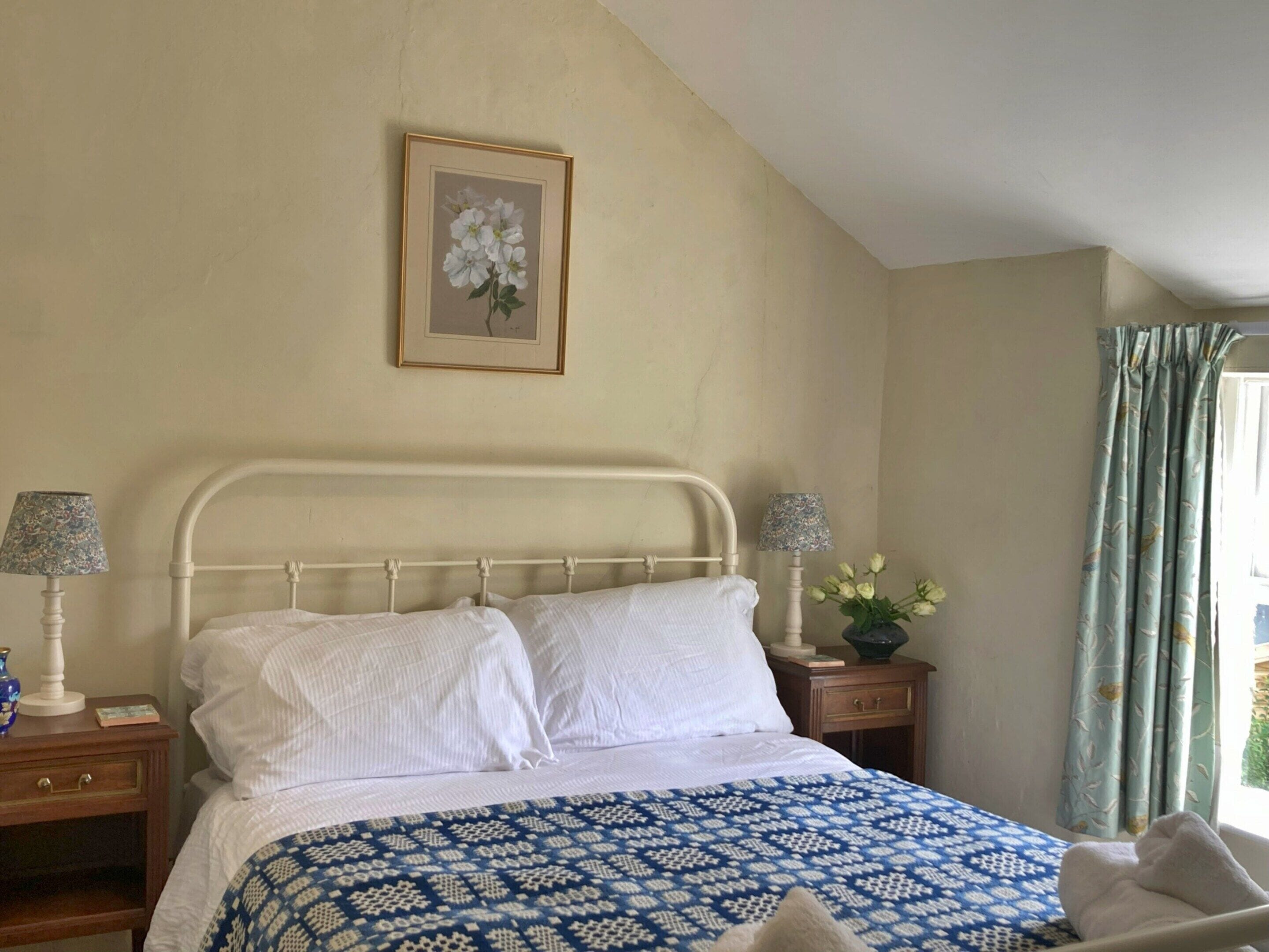 A bedroom with a bed, two bedside tables, a lamp, a framed picture on the wall, and a window with curtains.