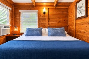 Een kamer met houten lambrisering, een bed, twee kussens, een wandlamp en een raam met zonwering.