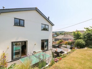Una casa moderna de dos pisos con techo plano, ventanas grandes y un patio con mesa y mobiliario exterior.