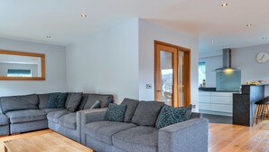 A modern living room with a sectional sofa, a wooden coffee table, and a kitchen area with a stove and a clock on the wall. - Scenic Lake View House for 8 (Torpoint)
