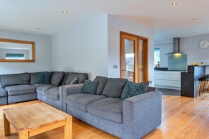 Ein modernes Wohnzimmer mit einem Ecksofa, einem Couchtisch aus Holz und einer Küchenzeile mit Herd und einer Wanduhr.