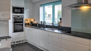 A modern kitchen with white cabinetry, a built-in oven, a microwave, and a stainless steel range hood. - Scenic Lake View House for 8 (Torpoint)