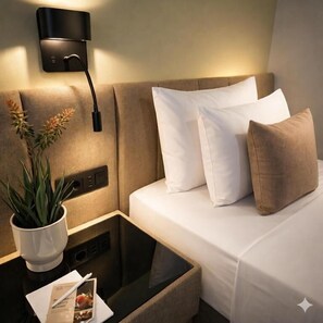 Habitación de hotel con cama, mesita de noche con lámpara, una planta y panel de control.