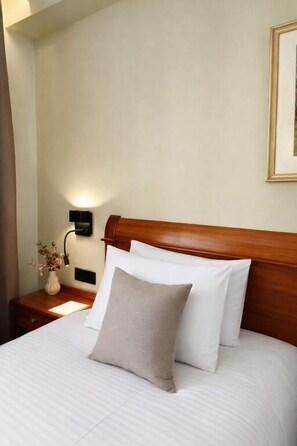 Een hotelkamer met een houten hoofdeinde, een bed met witte lakens, een bedlampje en een vaas met bloemen op het nachtkastje.