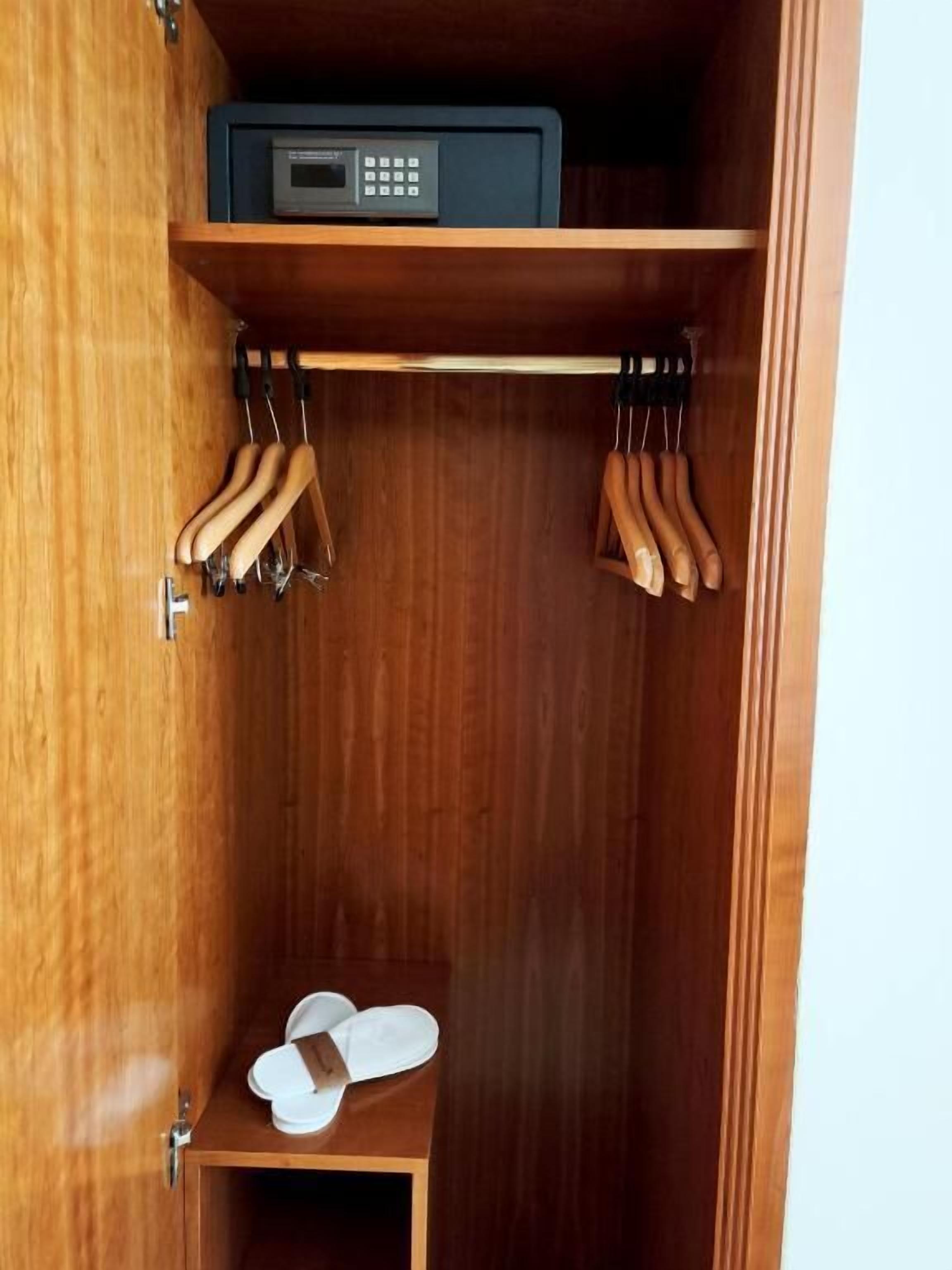 Un armario de madera con un estante que contiene un dispositivo electrónico negro, una barra para colgar con perchas de madera y un pequeño estante con zapatillas blancas.