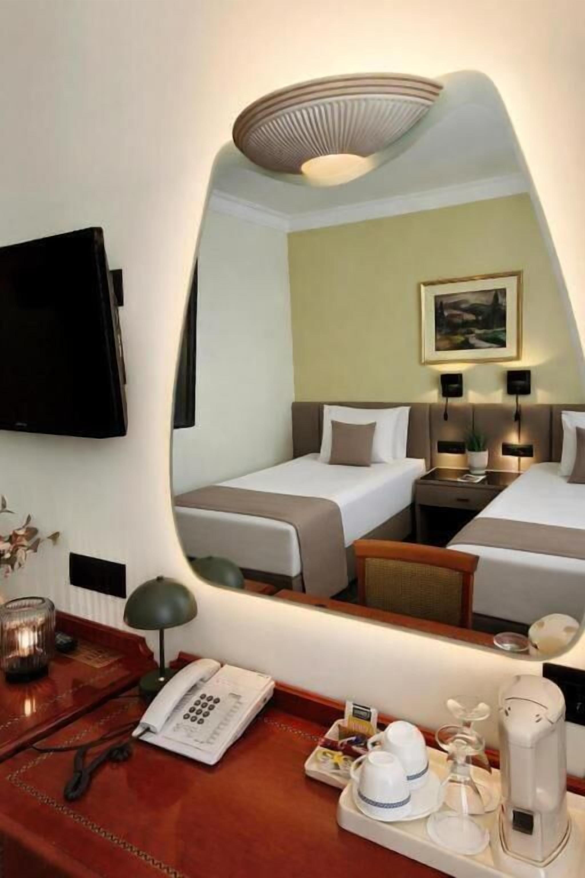 A hotel room with a bed, bedside table, lamp, and a mirror reflecting the room.