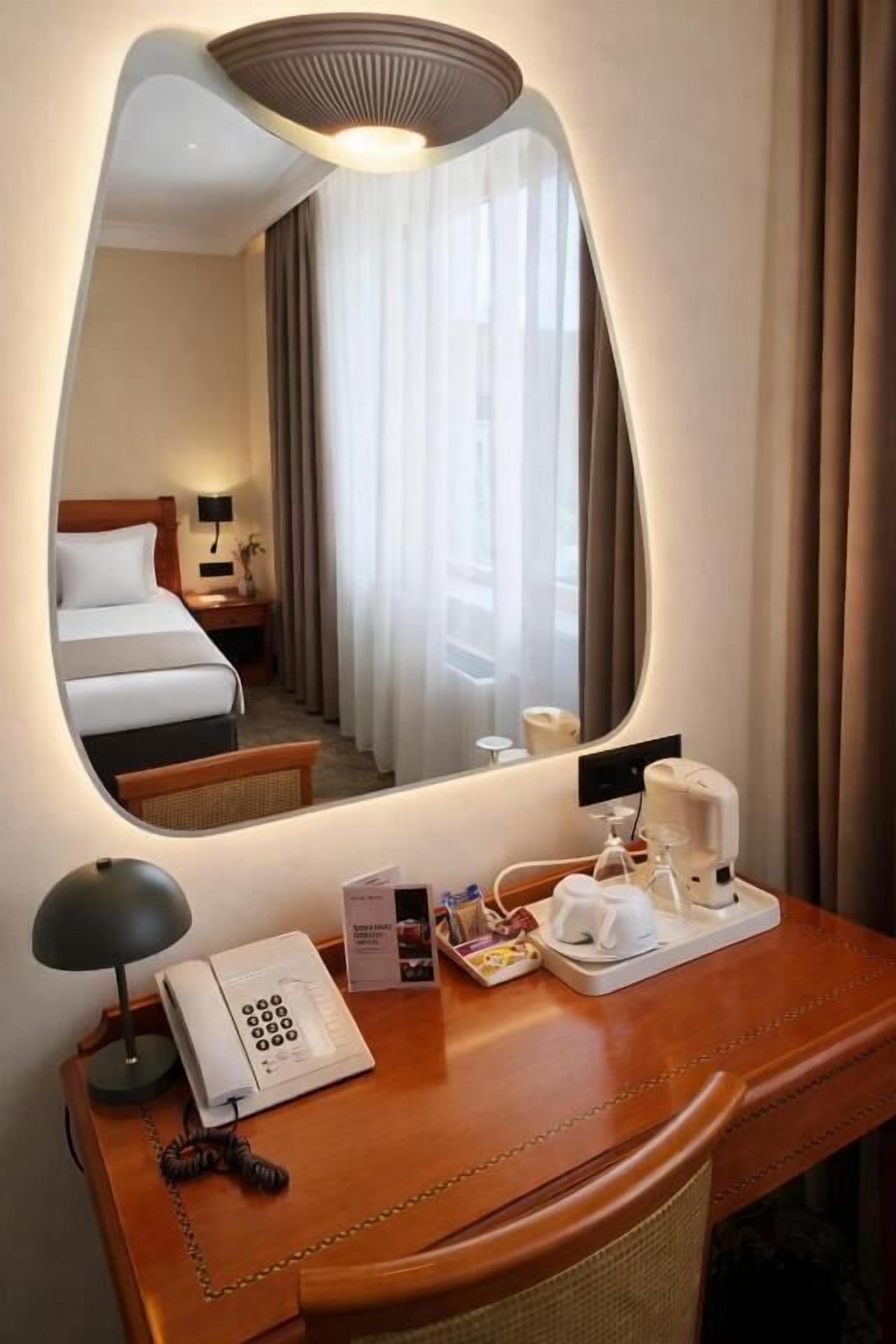 A hotel room with a large mirror, a bedside table with a telephone, a coffee maker, and a small table with a lamp and a book.
