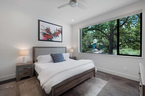 A bedroom with a bed, nightstands, a ceiling fan, a window with a view of trees, and a framed artwork on the wall. - Hot tub and fire pit deck near downtown (Austin)