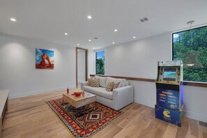 Ein modernes Wohnzimmer mit einem weißen Sofa, einem Couchtisch, einem farbenfrohen Teppich, einem Gemälde einer Naturlandschaft und einer Pac-Man Arcade-Maschine.