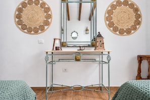 Una habitación con una mesa de vidrio, dos adornos tejidos en la pared, un espejo y un taburete de madera pequeño.