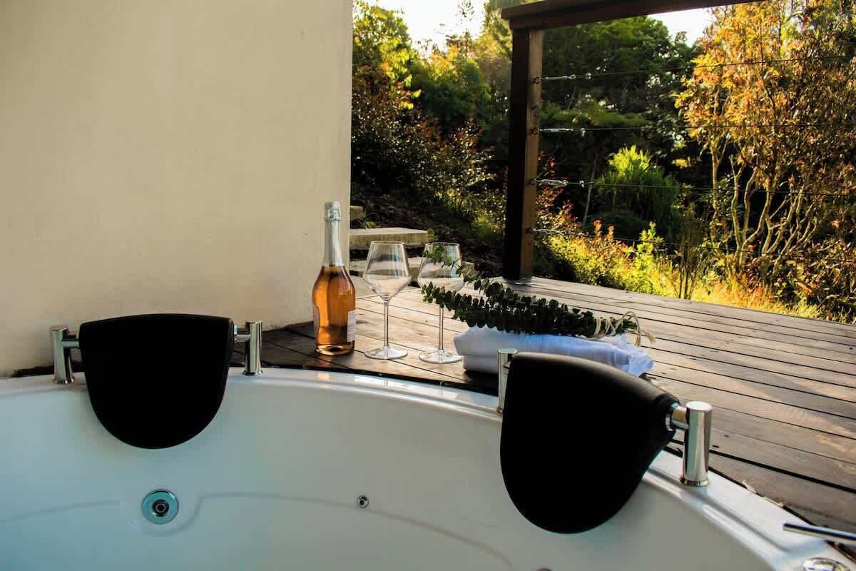 A hot tub with two black seats, a bottle of wine, and glasses on a wooden deck with a view of a garden.