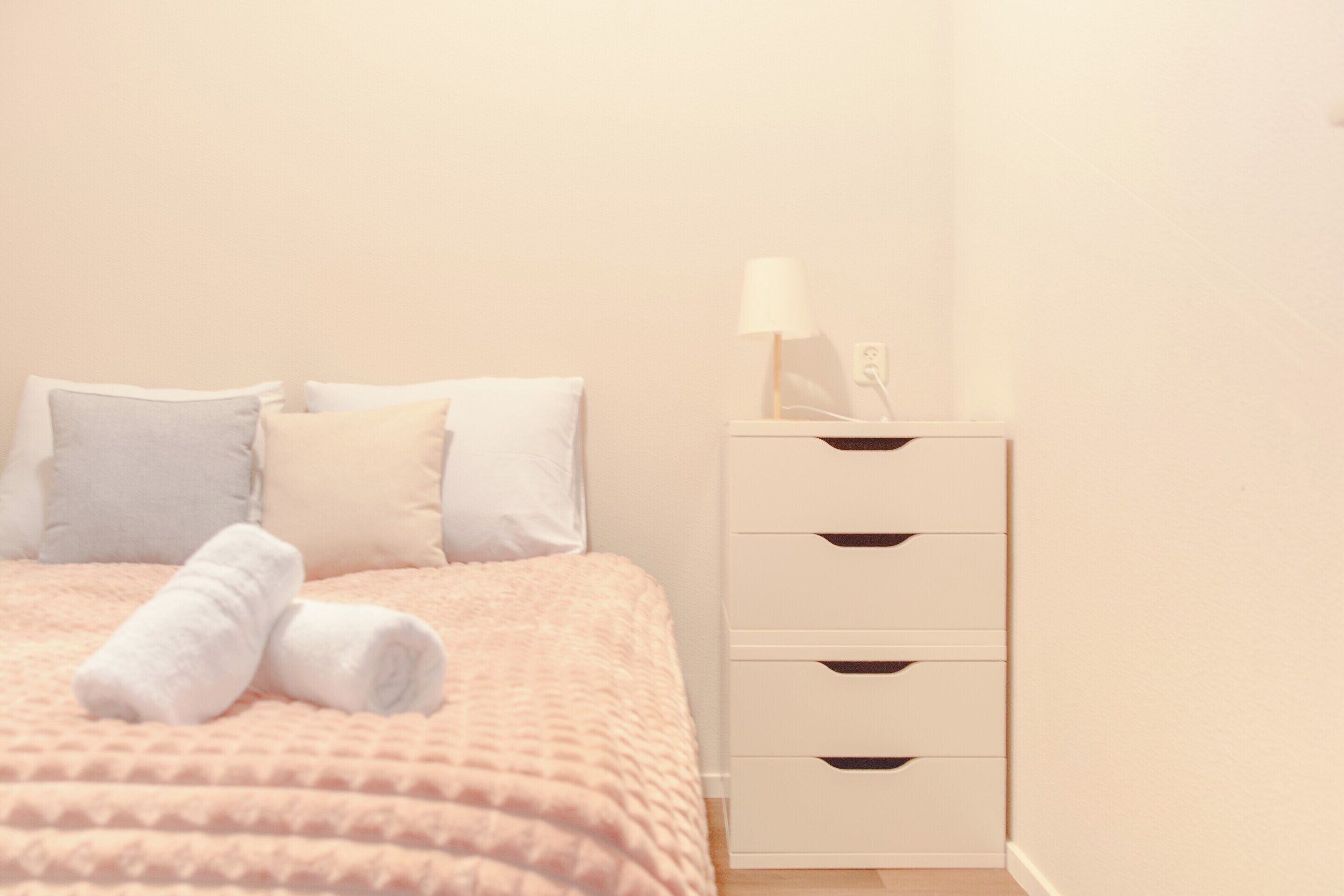 Een bed met een roze deken en witte kussens, een nachtkastje met een lamp, en een witte ladekast.