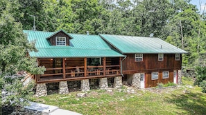 A log cabin with a green metal roof, stone foundation, and a porch with wooden furniture.
