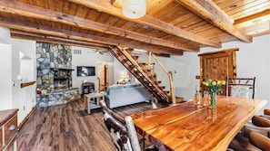 A wooden interior with a stone fireplace, a sofa, a dining table with chairs, and a wooden staircase.