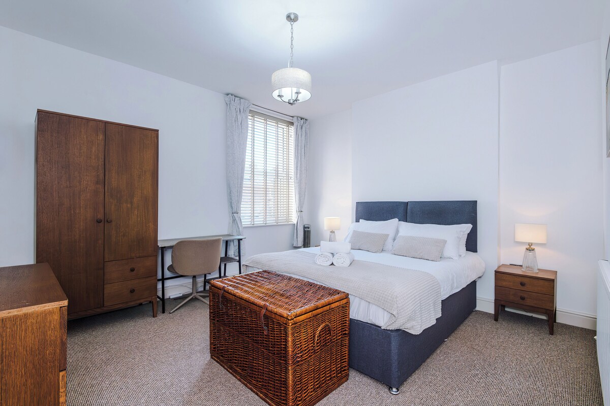 Ein Hotelzimmer mit Bett, Schreibtisch, Kleiderschrank, einem Korbstuhl-Nachttisch und einer Pendelleuchte.