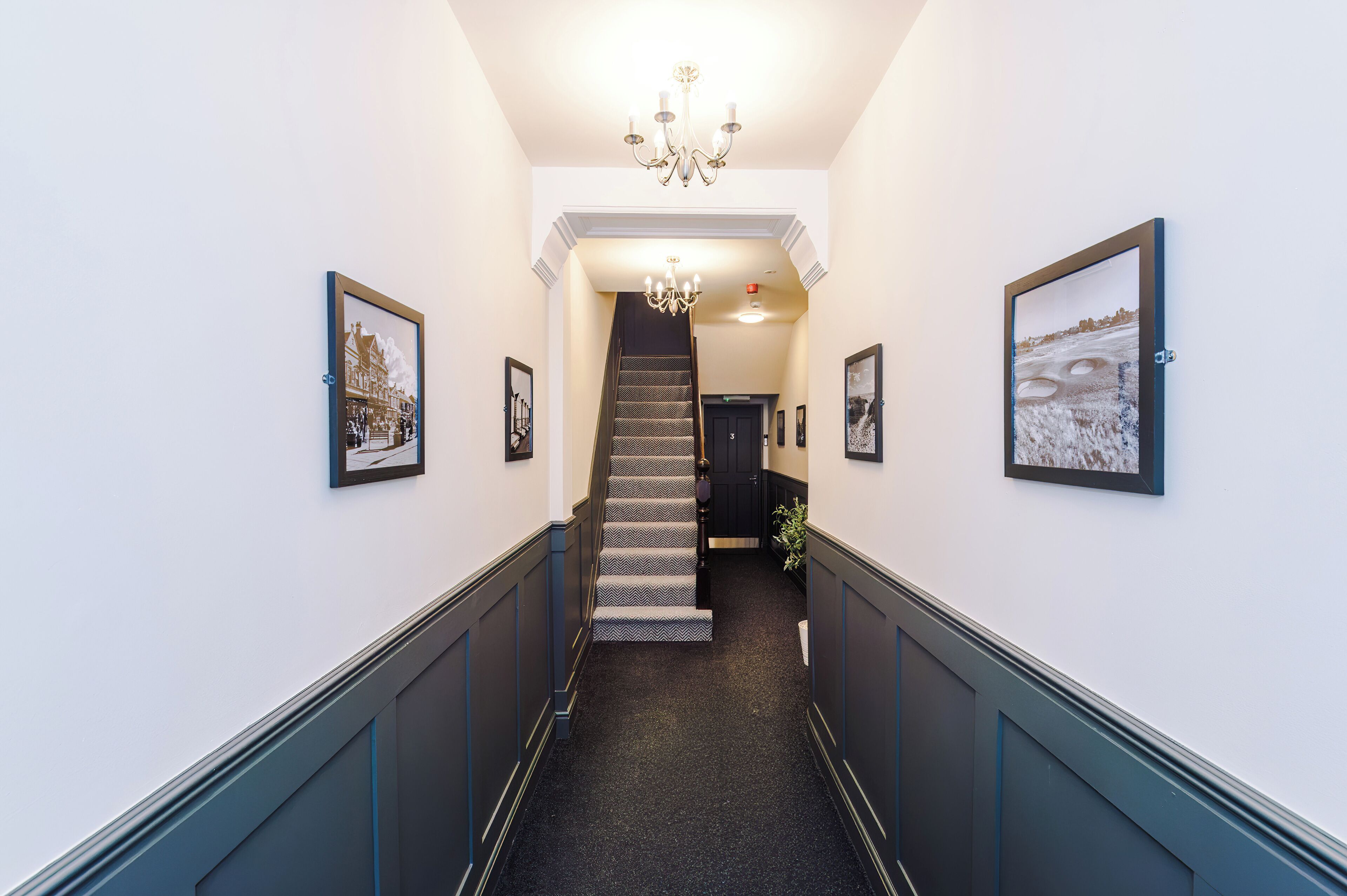 A hallway with a patterned carpet, a chandelier, and framed pictures on the walls.