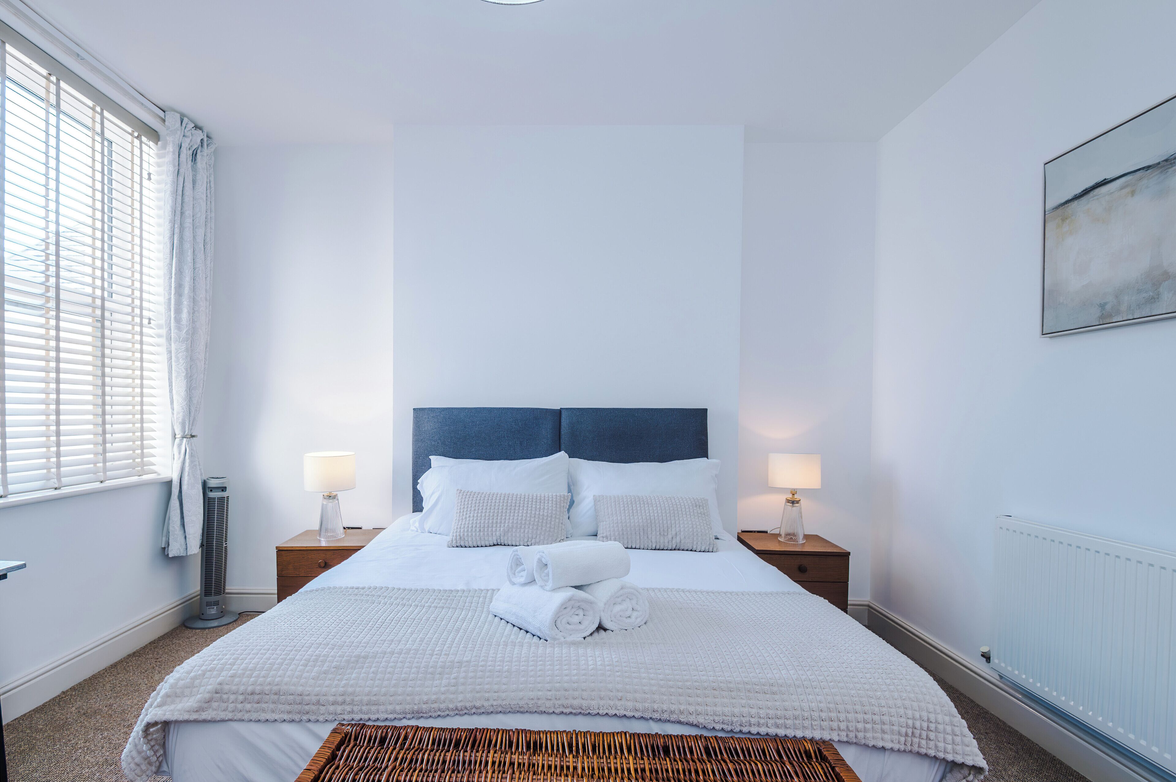 Habitación de hotel con una cama grande, dos mesitas de noche con lámparas, un radiador y una ventana con persianas.