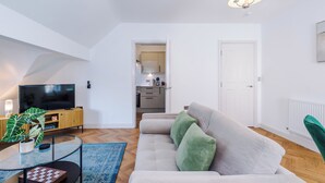 Een moderne woonkamer met een flatscreen televisie, een bank met groene kussens, een glazen salontafel, en een keuken op de achtergrond.