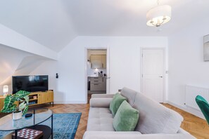Ein modernes Wohnzimmer mit einem Flachbildfernseher, einem Sofa mit grünen Kissen, einem Glastisch und einer Küche im Hintergrund.