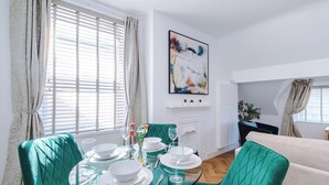 Een eethoek met een glazen tafel gedekt voor vier personen, groene bekleden stoelen en een witte open haard met daarboven een modern abstract schilderij.
