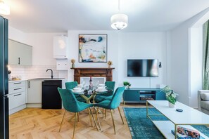A modern kitchen with a dishwasher, a dining area with a glass table and teal chairs, a flat-screen TV, and a fireplace. - The St Annes Beach Collection (Lytham St Annes)