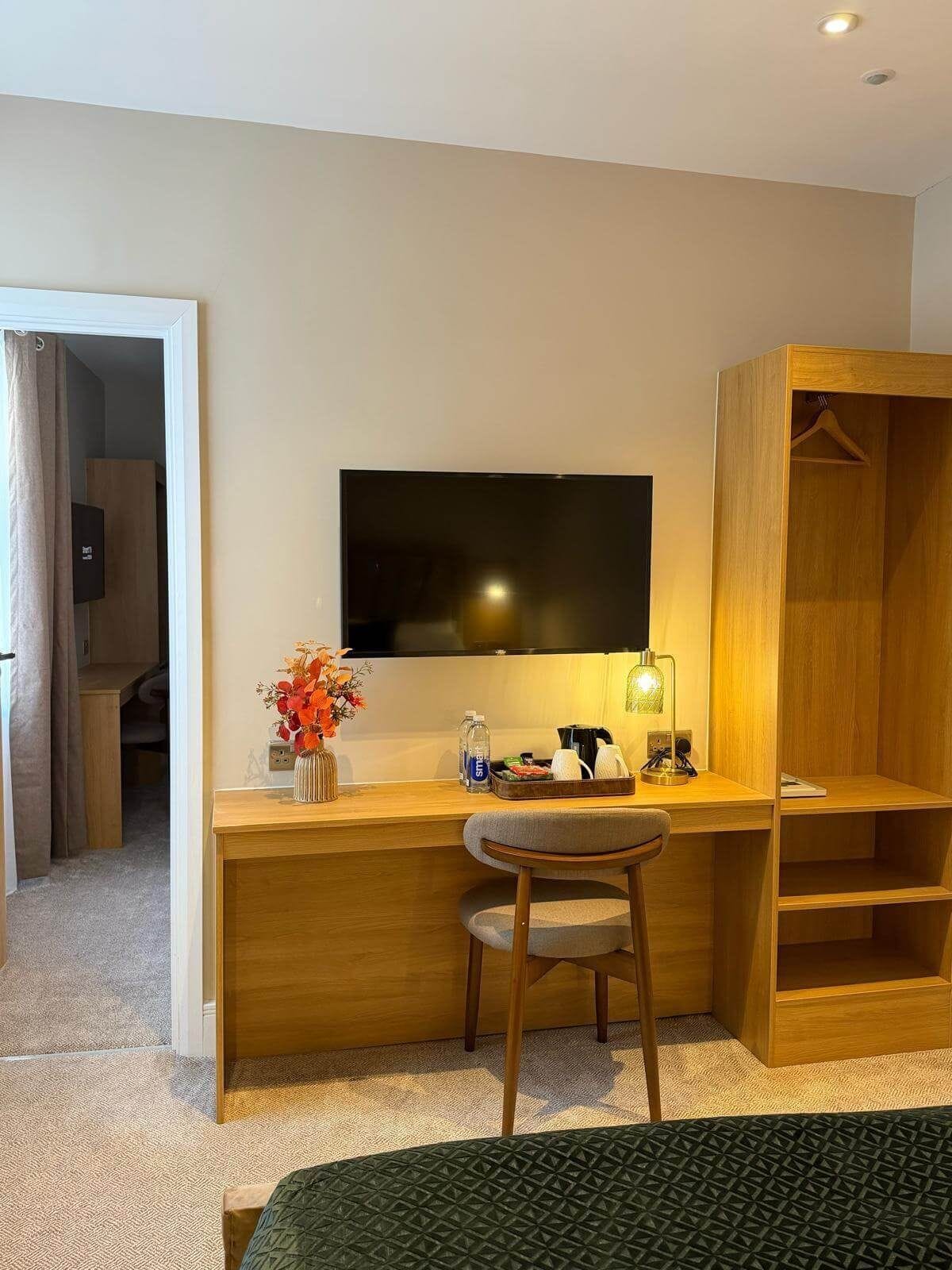 A hotel room with a wooden desk, a chair, a television mounted on the wall, and a wardrobe with a hanging space.