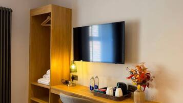 A hotel room with a wooden desk, a television mounted on the wall, a chair, a radiator, and a small plant.
