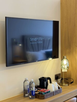 A flat-screen TV mounted on a wall, a coffee maker, two water bottles, a tray with various items, and a lamp on a wooden surface.