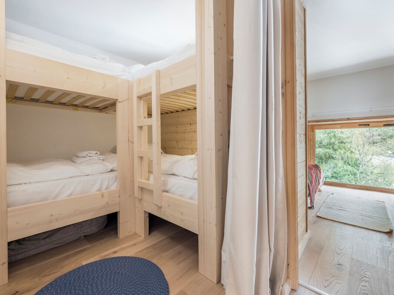 A room with two bunk beds, a window with a view of trees, and a blue rug on the floor.