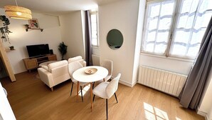 Uma sala de estar moderna com um sofá, uma mesa de jantar redonda e cadeiras. Há uma televisão em um suporte, uma planta e uma janela com cortinas.