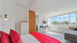 Moderni hotellihuone, jossa on sänky, työpöytä ja suuret ikkunat, joista avautuu näkymä kaupunkiin.
