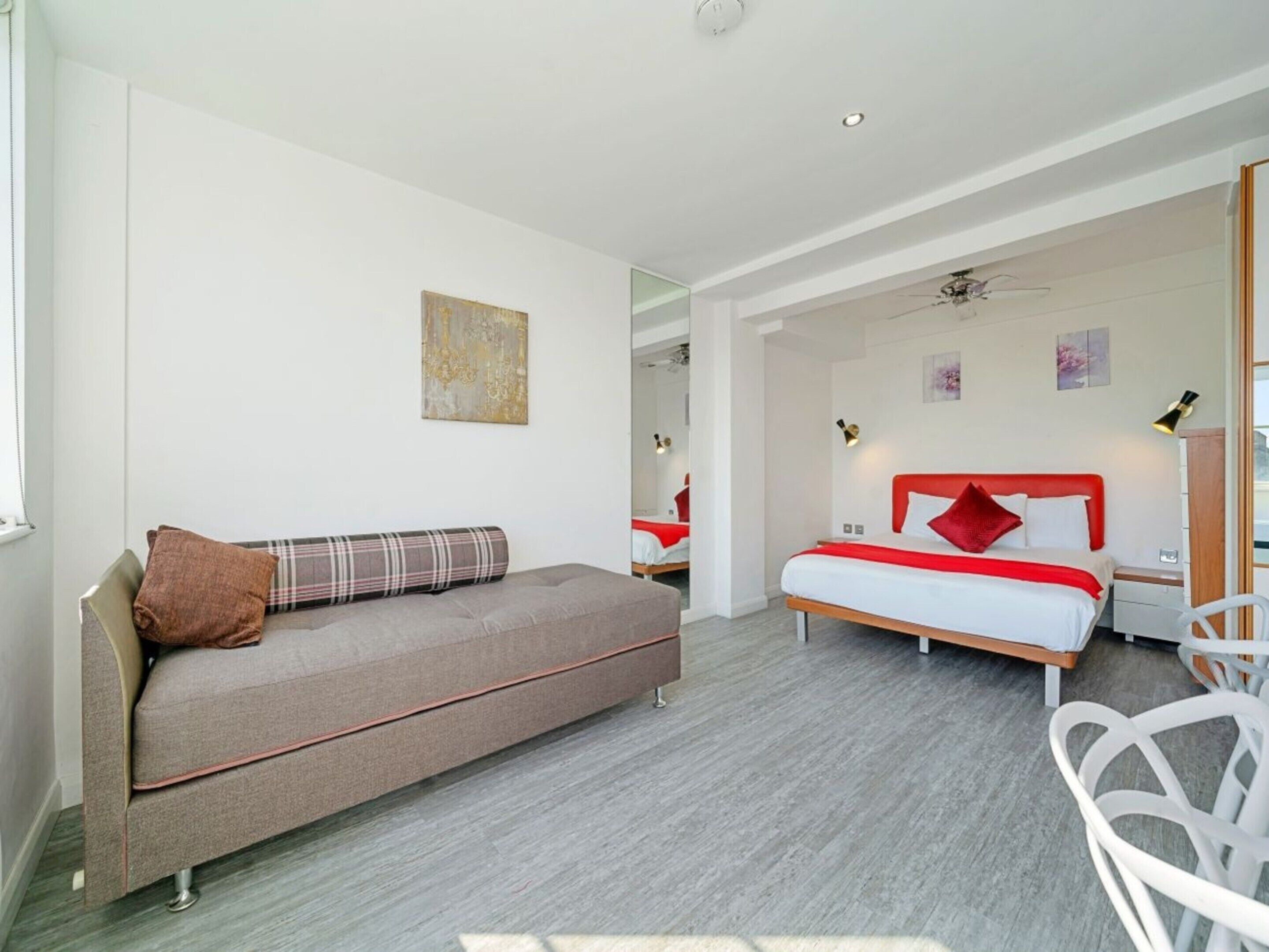 Moderni hotellihuone, jossa on sohva, punaisella vuodevaatteella varustettu sänky, peili ja kattotuuletin.