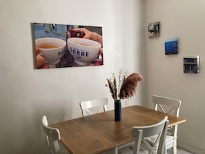 Uma área de refeições com uma mesa de madeira e cadeiras brancas, um vaso com plantas secas e uma parede com quadros.