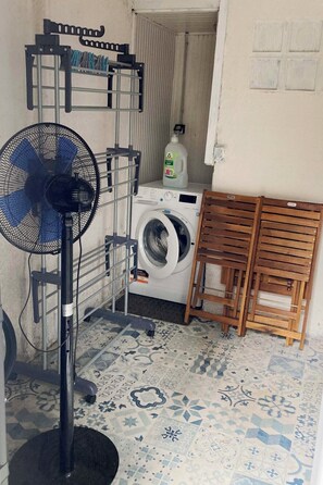 Ein Waschbereich mit Waschmaschine, Wäscheständer, Ventilator und einer Flasche Waschmittel.