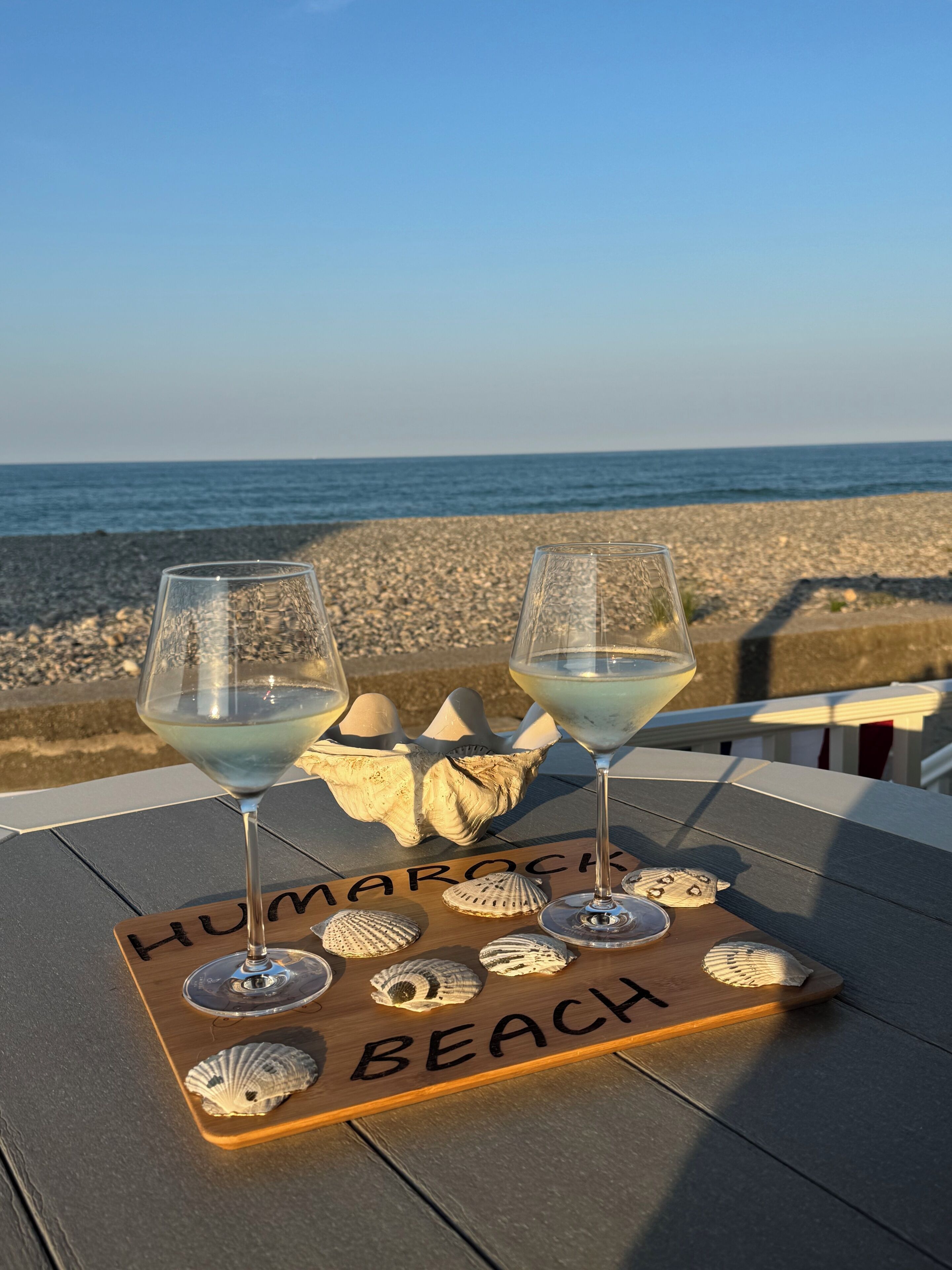 Two glasses of white wine on a wooden board with the words 'Humorrock Beach' and seashells.