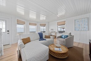 Ein geräumiges Wohnzimmer mit einer großen Couch, einem kleineren Sessel und einem runden Couchtisch. Mehrere Fenster mit Jalousien, Blick ins Freie und eine Wand mit dekorativem Muschelmuster.