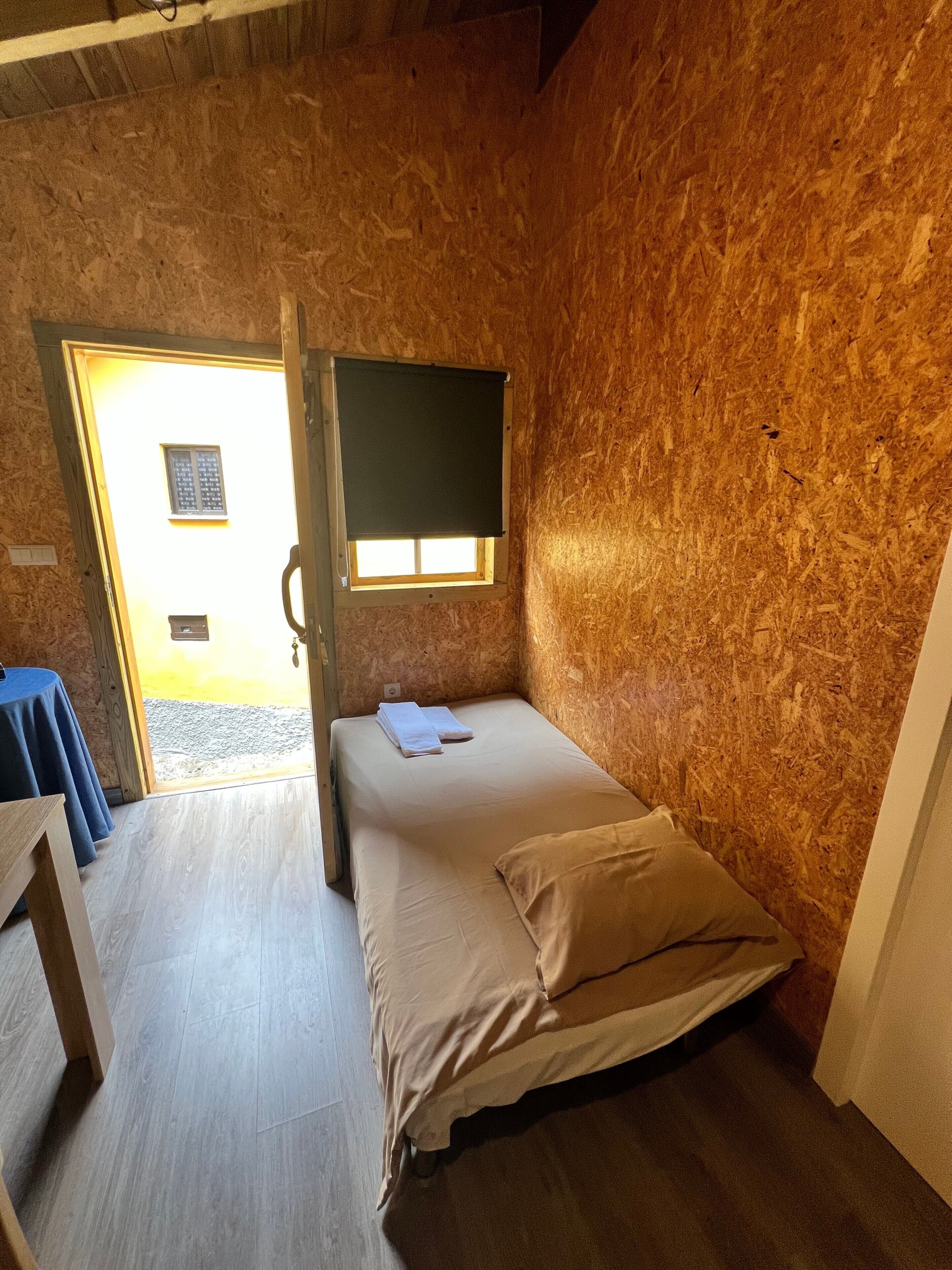Ein kleines Zimmer mit einem Einzelbett, einer Holzwand, einem Fenster mit Jalousie und einer Tür, die nach draußen führt.
