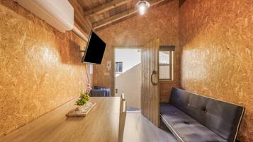 Une pièce avec un plafond en bois, un téléviseur fixé au mur, un banc, une petite table avec une plante et une porte donnant sur une autre pièce.