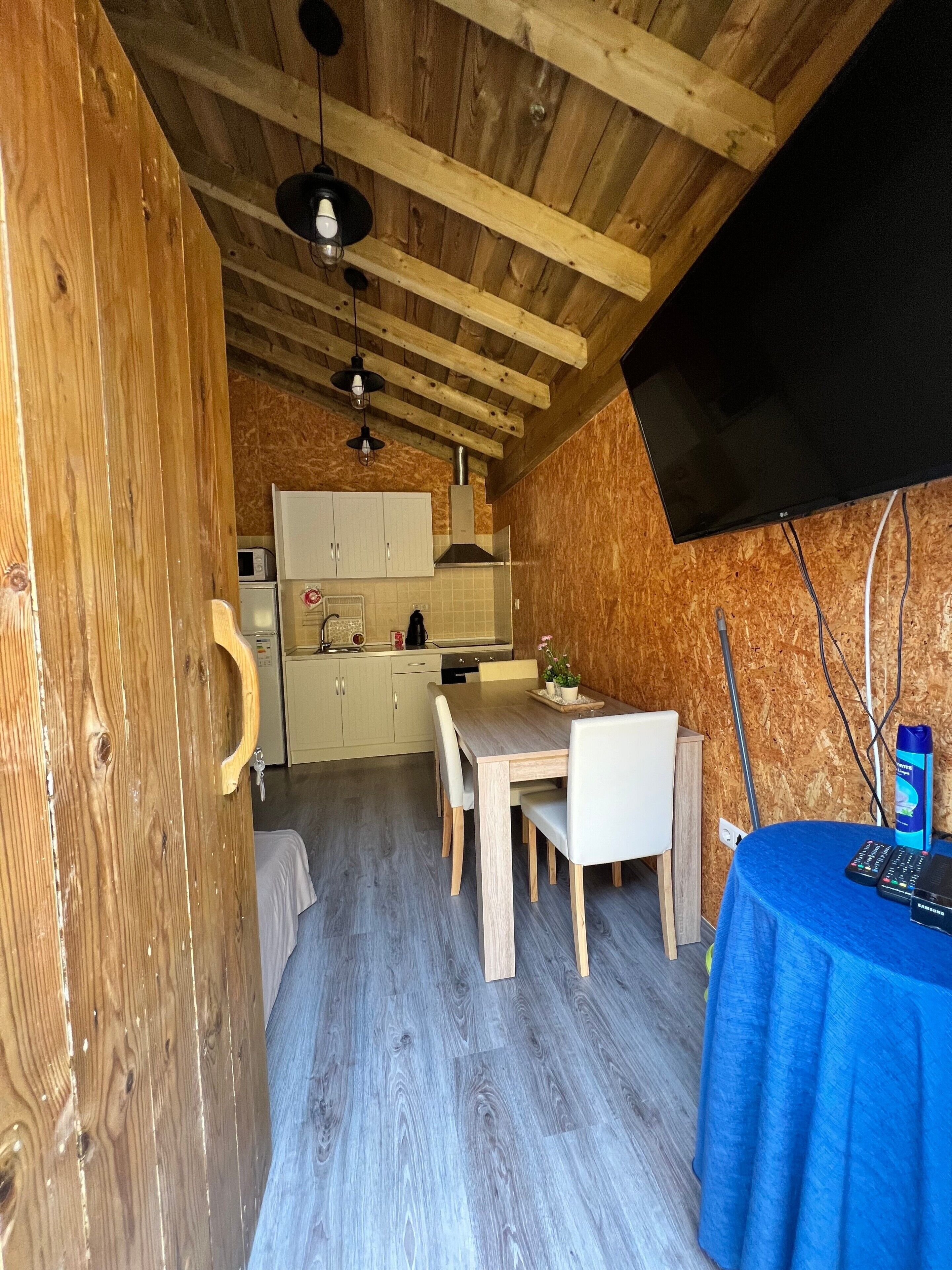 A compact kitchen with wooden beams, a dining table, and a flat-screen TV mounted on the wall.