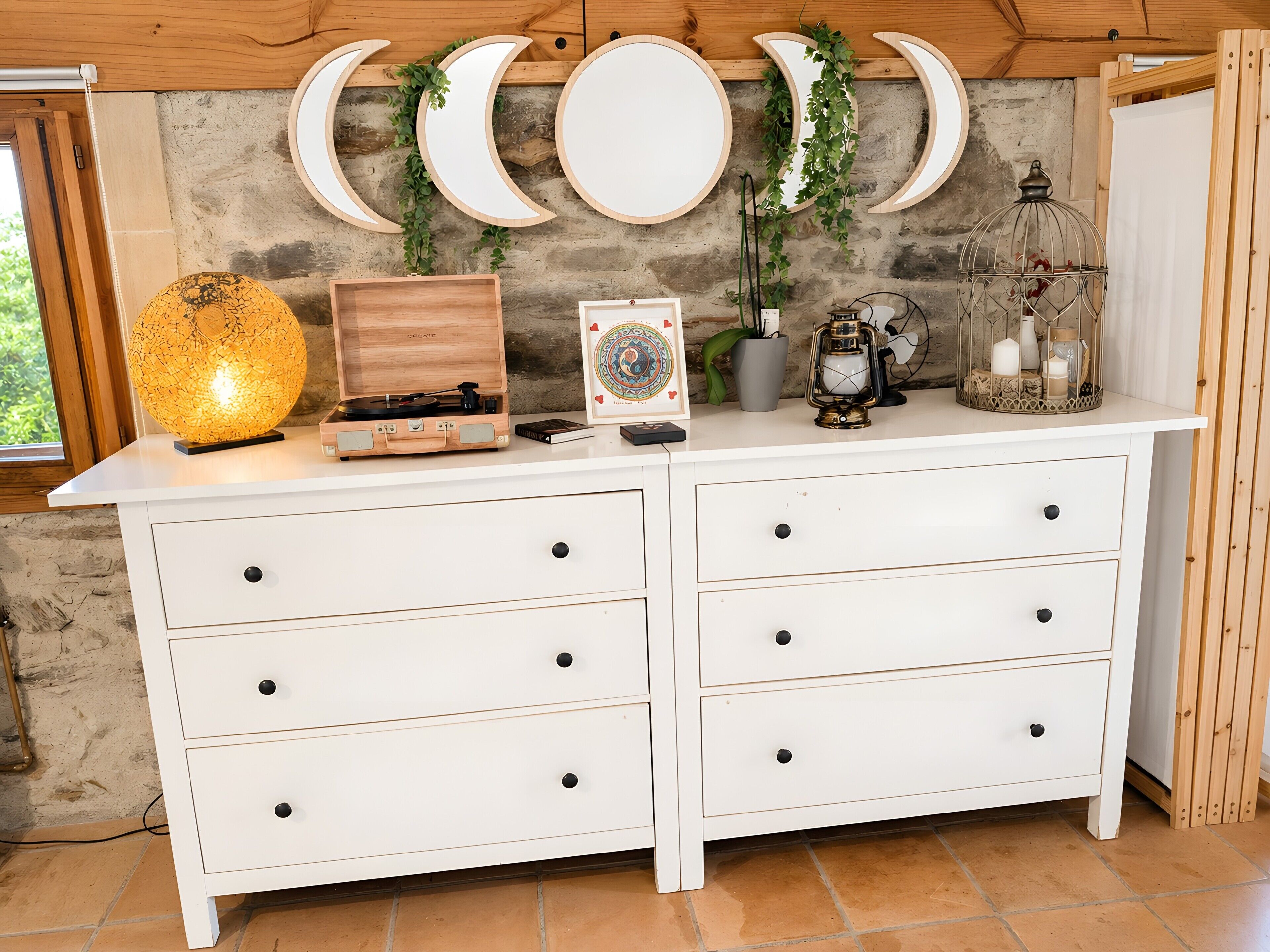 Een witte dressoir met meerdere lades, een houten platenspeler, een ingelijst beeld, een lantaarn en wanddecoraties in de vorm van een halve maan.